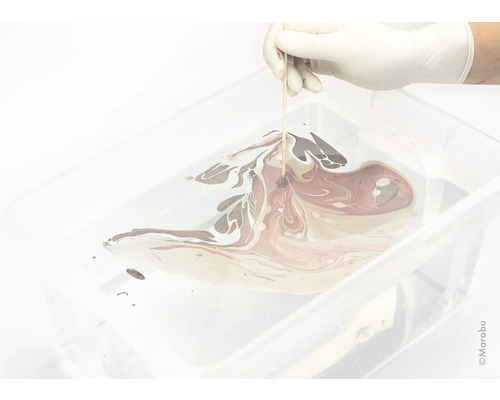 Een hand met een handschoen marmert verf in een bak met een houten stok.