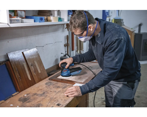 Een man schuurt een stuk hout met een excentrische schuurmachine in een werkplaats