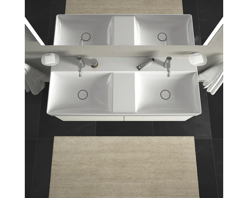 Bovenaanzicht van een dubbele wastafel met kranen, zeepbakjes, handdoeken en een vloerkleed in de badkamer.