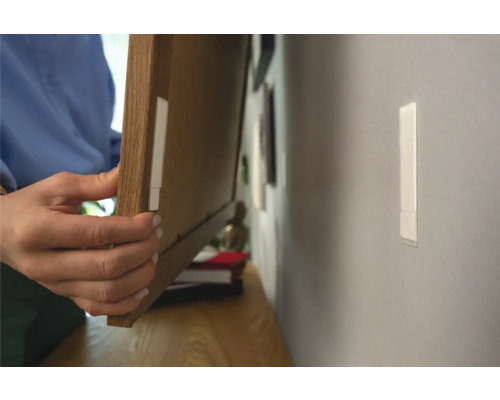 Persoon bevestigt fotolijst met plakstrips aan de muur.