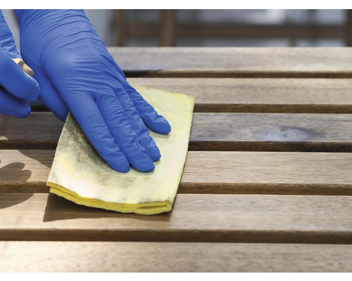 Een hand met beschermende handschoen reinigt een houten oppervlak met een doek.