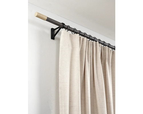 Donkere gordijnroede van hout en metaal met lichte eindkappen, zwarte ringen en beige gordijn aan een zwarte wandsteun.