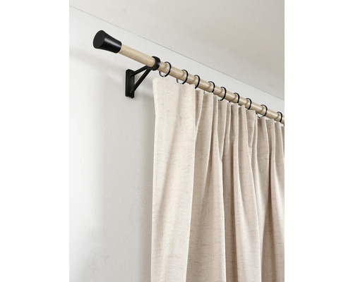 Houten gordijnroede met zwarte eindknoppen en ringen en een beige gordijn met structuur aan de wand.