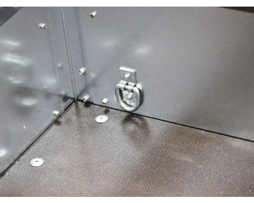 Metalen sjoroog aan de zijwand van een aanhangwagen voor ladingzekering boven een gestructureerde vloer.