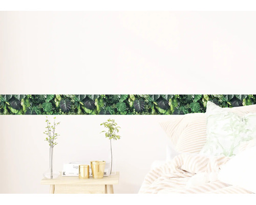 Zelfklevende behangrand met tropisch monstera bladerpatroon in een lichte slaapkamer boven een bed.