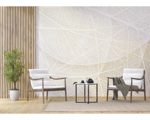 Lichte woonkamer met fotobehang in bladnerf-design, twee houten fauteuils met witte kussens, zwarte bijzetmeter en kamerplant.