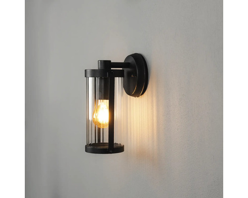 Moderne buitenwandlamp met zwart metalen frame, cilindrische glazen kap en brandende lichtbron.