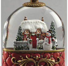 Sneeuwbol kerstdecoratie met besneeuwd huis, sneeuwpop en dennenbomen op een rode voet met gouden details.