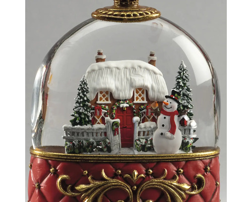 Sneeuwbol kerstdecoratie met besneeuwd huis, sneeuwpop en dennenbomen op een rode voet met gouden details.