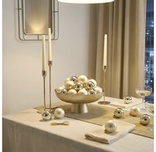 Feestelijk gedekte tafel met een schaal op voet vol kerstballen, hoge kandelaars en servetten in een lichte en elegante kamer.