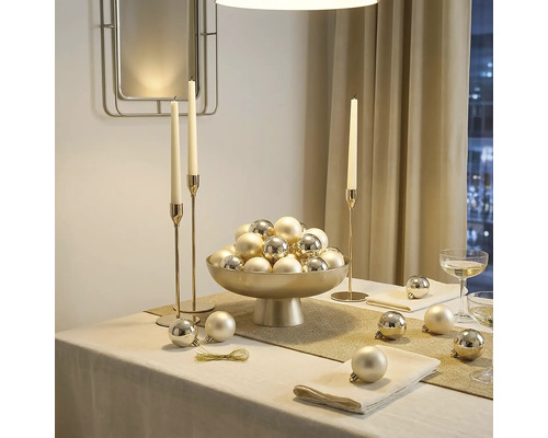 Feestelijk gedekte tafel met een schaal op voet vol kerstballen, hoge kandelaars en servetten in een lichte en elegante kamer.