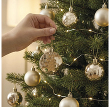Een hand hangt een gouden kerstbal in een groene kerstboom die is versierd met lampjes en kerstdecoratie.