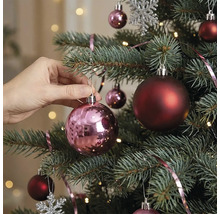 Een hand hangt een glanzende roze kerstbal in een versierde kerstboom met donkerrode ballen en lichtjes.