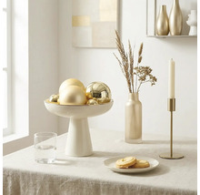 Heldere tafelsetting met een witte schaal op voet met gouden kerstballen, een slanke vaas met droogbloemen en een gouden kandelaar.