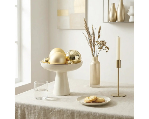 Heldere tafelsetting met een witte schaal op voet met gouden kerstballen, een slanke vaas met droogbloemen en een gouden kandelaar.