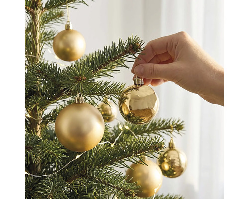 Een hand hangt een glanzende gouden kerstbal in een kerstboom met matte en glanzende kerstballen.