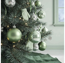 Kerstboom versierd met groene kerstballen, zilveren discoballen, kleine wanten en een lichtsnoer in een lichte kamer.