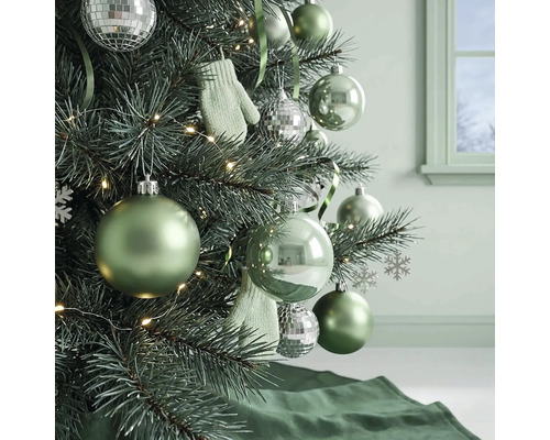 Kerstboom versierd met groene kerstballen, zilveren discoballen, kleine wanten en een lichtsnoer in een lichte kamer.