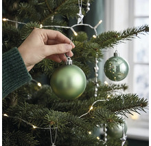 Een hand hangt een matgroene kerstbal in een versierde kerstboom met led-lichtsnoer.