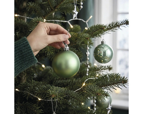 Een hand hangt een matgroene kerstbal in een versierde kerstboom met led-lichtsnoer.