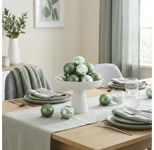 Feestelijk gedekte houten tafel met groene kerstballen in een witte schaal, servies, tafelloper en kaars in een lichte kamer.
