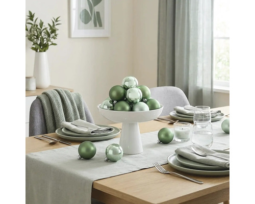 Feestelijk gedekte houten tafel met groene kerstballen in een witte schaal, servies, tafelloper en kaars in een lichte kamer.