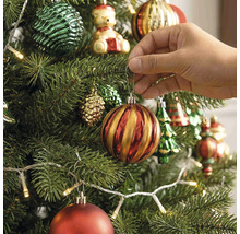 Een hand hangt een gestreepte kerstbal aan een versierde kerstboom met kerstverlichting en verschillende ornamenten.