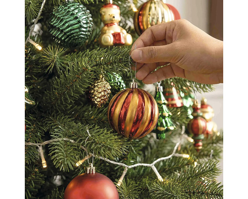 Een hand hangt een gestreepte kerstbal aan een versierde kerstboom met kerstverlichting en verschillende ornamenten.