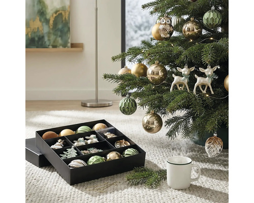 Kerstboom versierd met kerstballen, uilen- en hertfiguren naast een opbergdoos met kerstversiering op een tapijt.