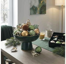 Feestelijke tafeldecoratie met een donkergroene schaal op voet gevuld met kerstversiering, een kaars en dennentakken op een houten tafel.