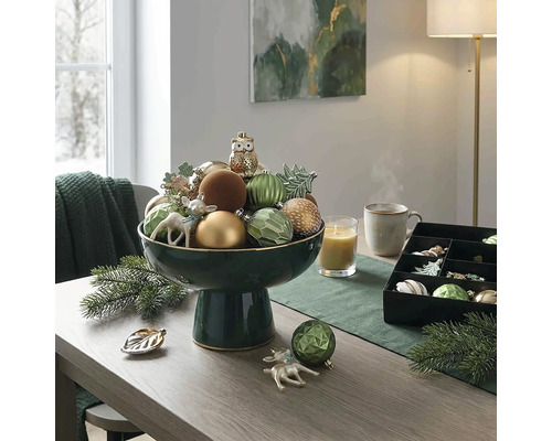 Feestelijke tafeldecoratie met een donkergroene schaal op voet gevuld met kerstversiering, een kaars en dennentakken op een houten tafel.