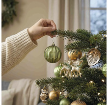 Hand hangt een groene geometrische kerstbal in een versierde kerstboom met herten- en uilenfiguren en warme verlichting.