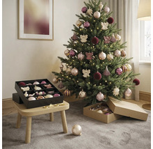 Versierde kerstboom in lichte kamer met warmwit licht, ballen in roze en paars en een zwarte opbergdoos op een houten krukje.