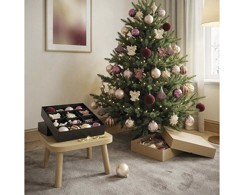 Versierde kerstboom in lichte kamer met warmwit licht, ballen in roze en paars en een zwarte opbergdoos op een houten krukje.