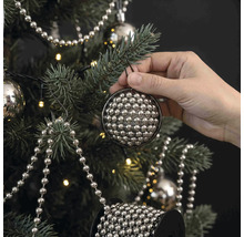 Een hand hangt een ronde kerstbal van kleine zilveren kraaltjes in een versierde kerstboom met lichtsnoer en parelslingers.