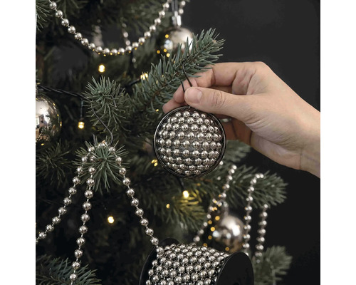Een hand hangt een ronde kerstbal van kleine zilveren kraaltjes in een versierde kerstboom met lichtsnoer en parelslingers.