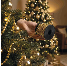 Hand houdt rol parelslinger vast bij het versieren van een kerstboom met kerstballen en verlichting.