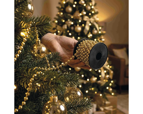 Hand houdt rol parelslinger vast bij het versieren van een kerstboom met kerstballen en verlichting.