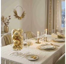Feestelijk gedekte eettafel met gouden kerstballen in een glazen cilinder, kaarsen, goudkleurig bestek en elegant servies.