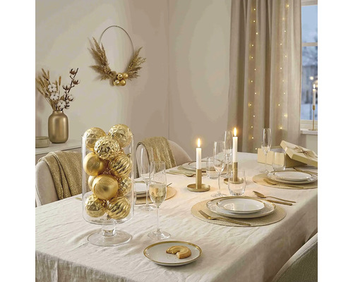 Feestelijk gedekte eettafel met gouden kerstballen in een glazen cilinder, kaarsen, goudkleurig bestek en elegant servies.
