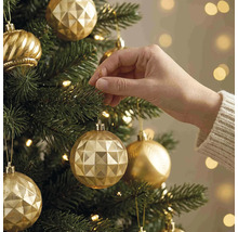 Een hand hangt een gouden kerstbal met geometrisch patroon in een groene kerstboom met warme lichtjes.