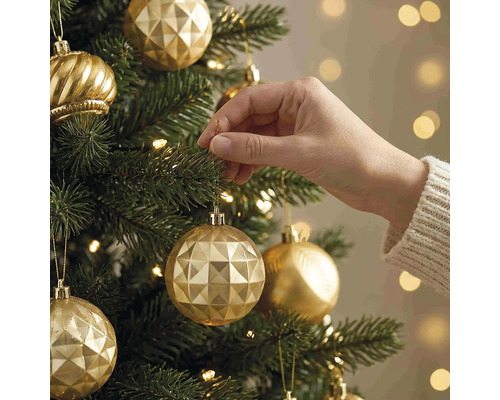 Een hand hangt een gouden kerstbal met geometrisch patroon in een groene kerstboom met warme lichtjes.
