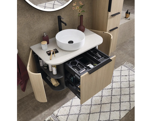 Modern badkamermeubel in houtlook met witte opzetwastafel, zwarte kraan en open lade met vakverdeling.