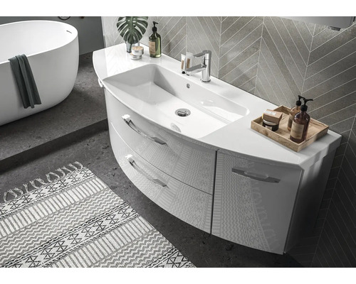 Moderne, lichte badkamer met gebogen wastafelmeubel, twee laden en kraan. In de achtergrond een vrijstaand ligbad en wandtegels.