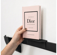 Een hand plaatst een boek op een minimalistische, zwarte metalen wandplank aan een muur.