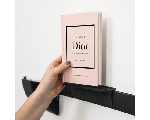 Een hand plaatst een boek op een minimalistische, zwarte metalen wandplank aan een muur.