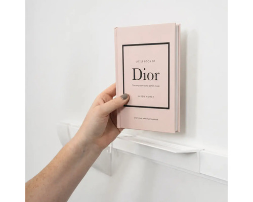Een hand plaatst het boek Little Book of Dior op een smalle witte wandplank.