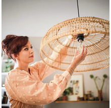 Een vrouw vervangt een lichtbron in een ronde hanglamp van gevlochten rotan in een sfeervol ingerichte kamer.