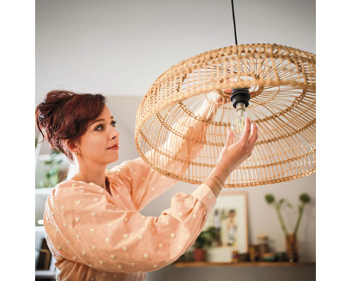Een vrouw vervangt een lichtbron in een ronde hanglamp van gevlochten rotan in een sfeervol ingerichte kamer.