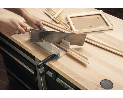 Iemand zaagt een fotolijst met een handzaag en verstekbak op een werkbank.